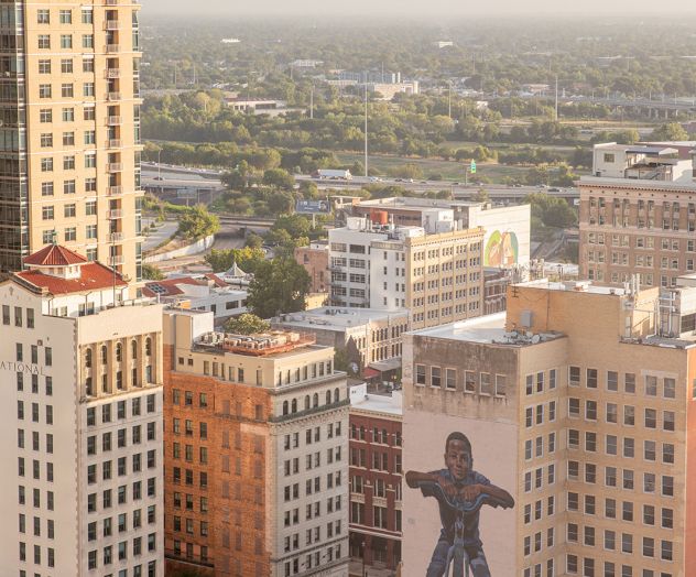 An aerial view of a cityscape with a notable building mural featuring a large figure, surrounded by tall buildings and greenery.