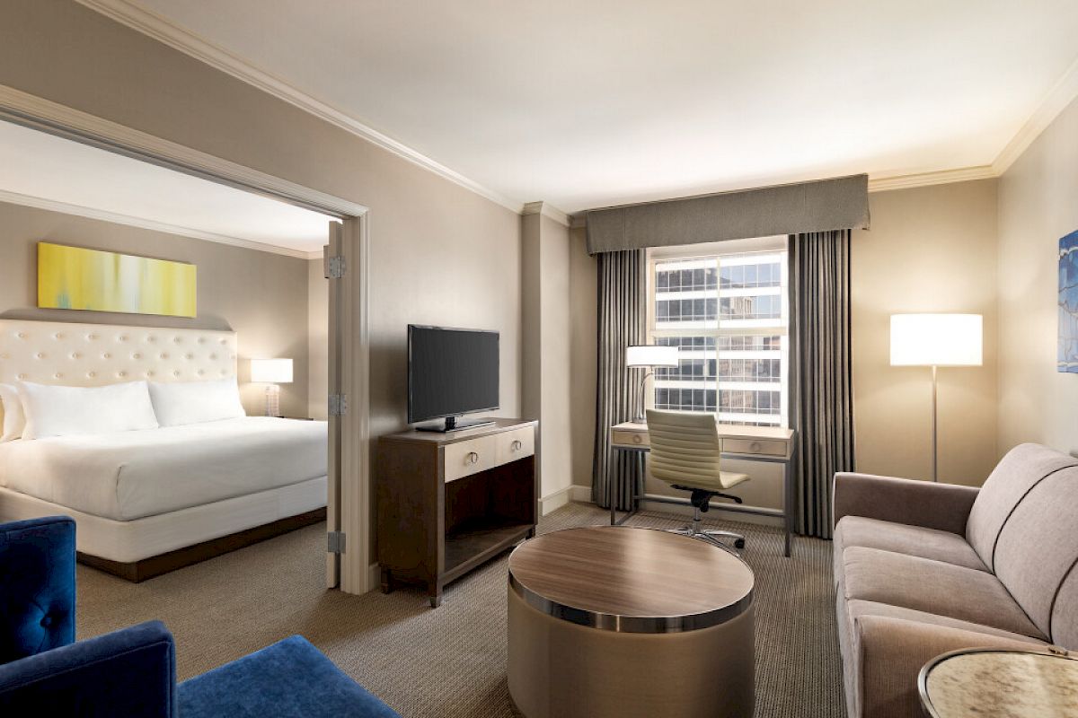 The image shows a hotel suite with a bedroom, living area, TV, sofa, desk, and window with a city view. The decor is modern and neutral.