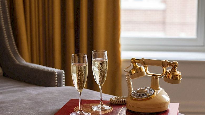 A luxurious setting with two filled champagne glasses and a vintage phone on a tray by a window with yellow curtains.