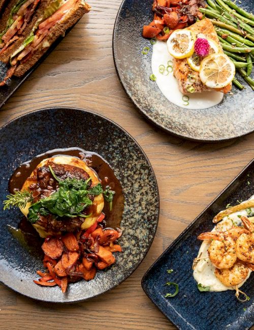 A table with four gourmet dishes, including fries, steak, seafood, and salmon, with a menu visible in the corner on a wooden surface.