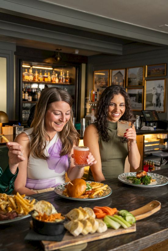 Two people enjoying drinks and a variety of food at a restaurant table, featuring fries, veggies, and more delicious dishes.