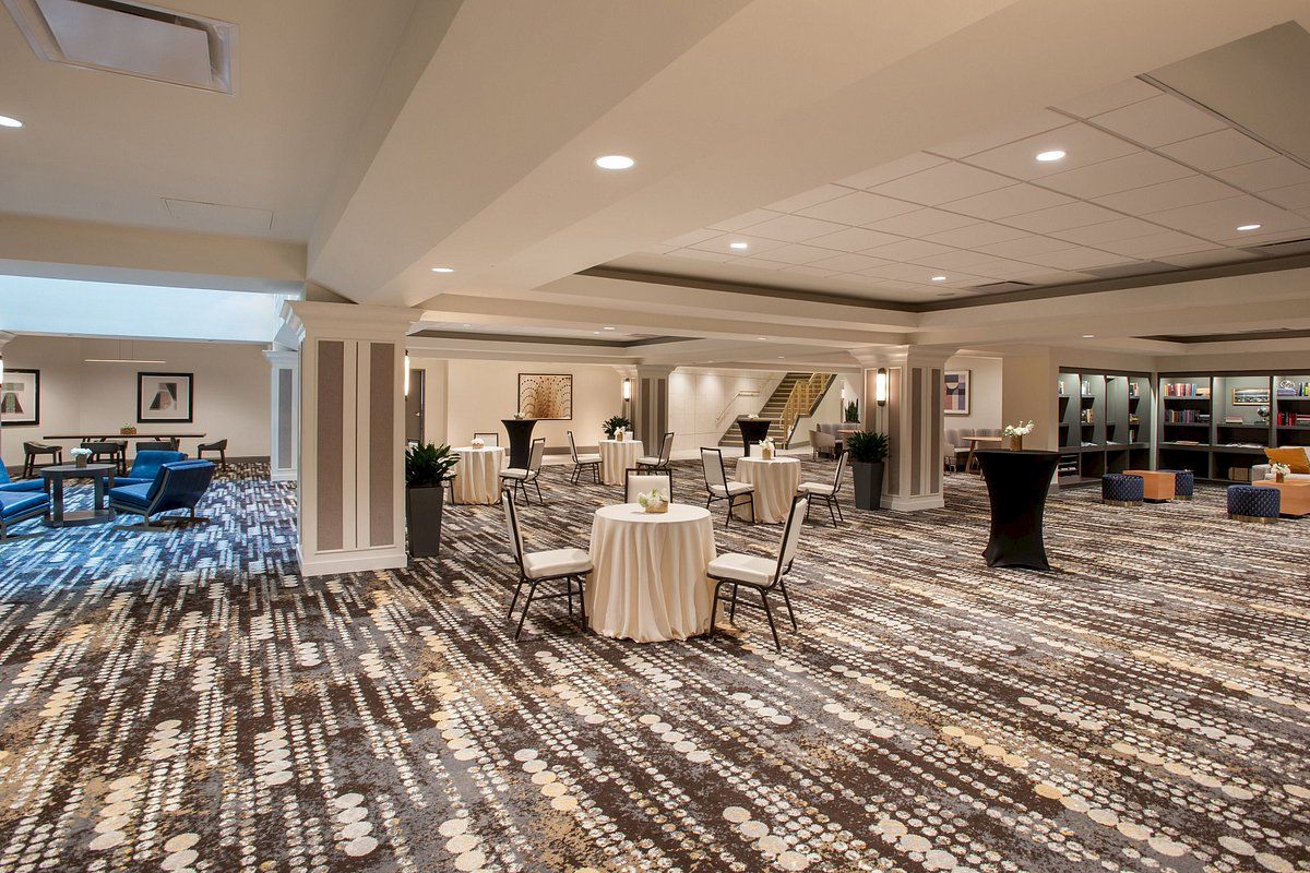 The image shows a spacious room with round tables and chairs, elegant carpeting, and modern decor, likely set up for an event.