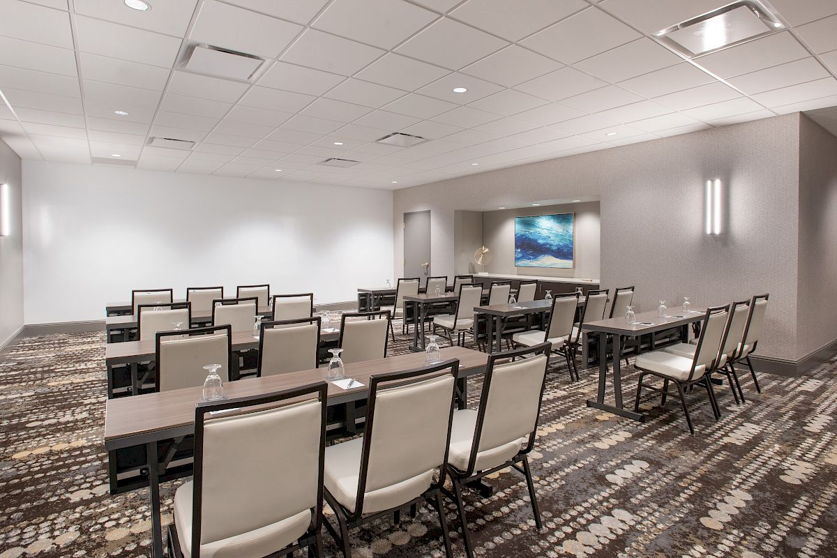 The image shows a modern conference room with rows of chairs and tables, a patterned carpet, and a painting on the wall.