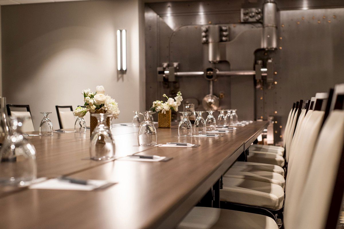 The image shows a long conference table set with upside-down glasses, notepads, and flowers, in a room with industrial decor elements.