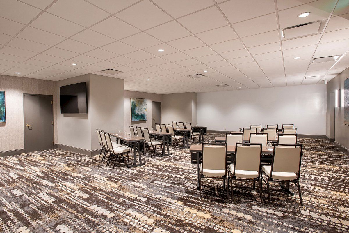 The image shows a conference room with rows of chairs and tables on a patterned carpet. There's a TV on the wall and framed artwork.