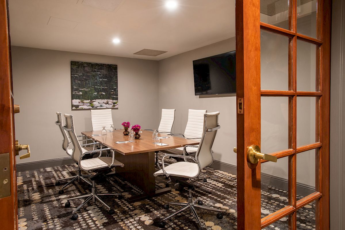 A modern conference room with a wooden table, white chairs, a wall-mounted TV, and artwork. The room has soft lighting and a patterned carpet.