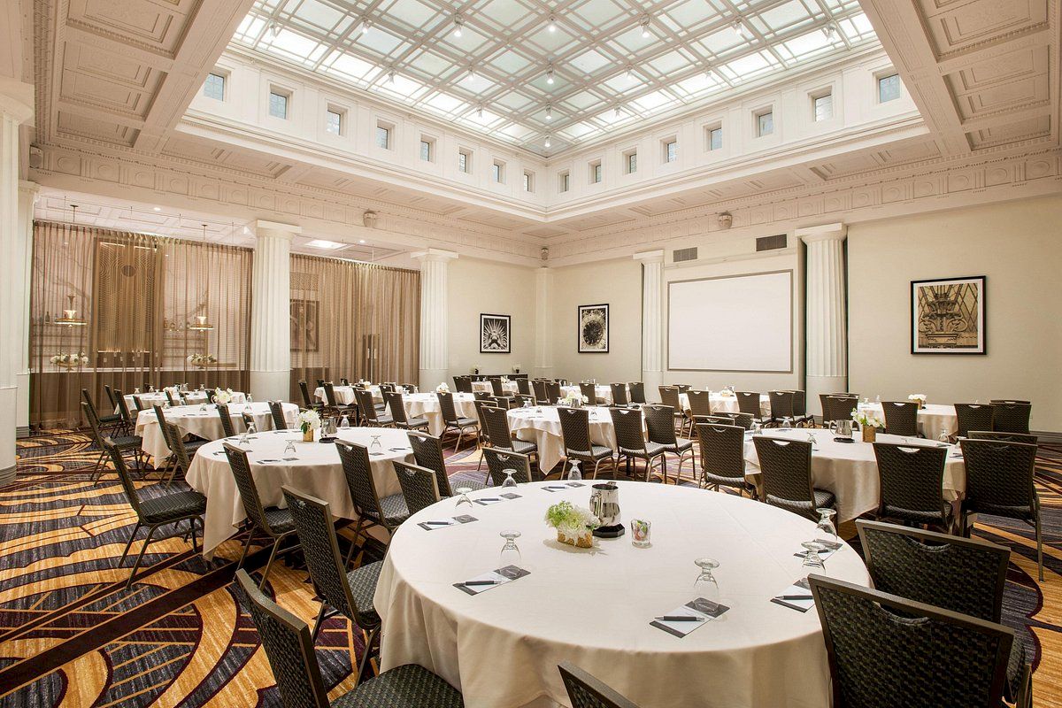 The image shows a conference room with round tables, chairs, a large screen, and high ceiling. The room is set up for a meeting or event.