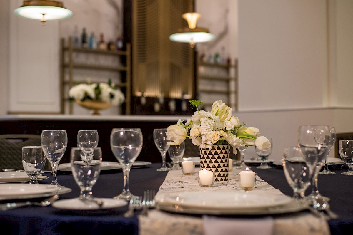 A dining table set elegantly with glassware, plates, and a floral centerpiece, in a room with soft lighting and a decorative background.