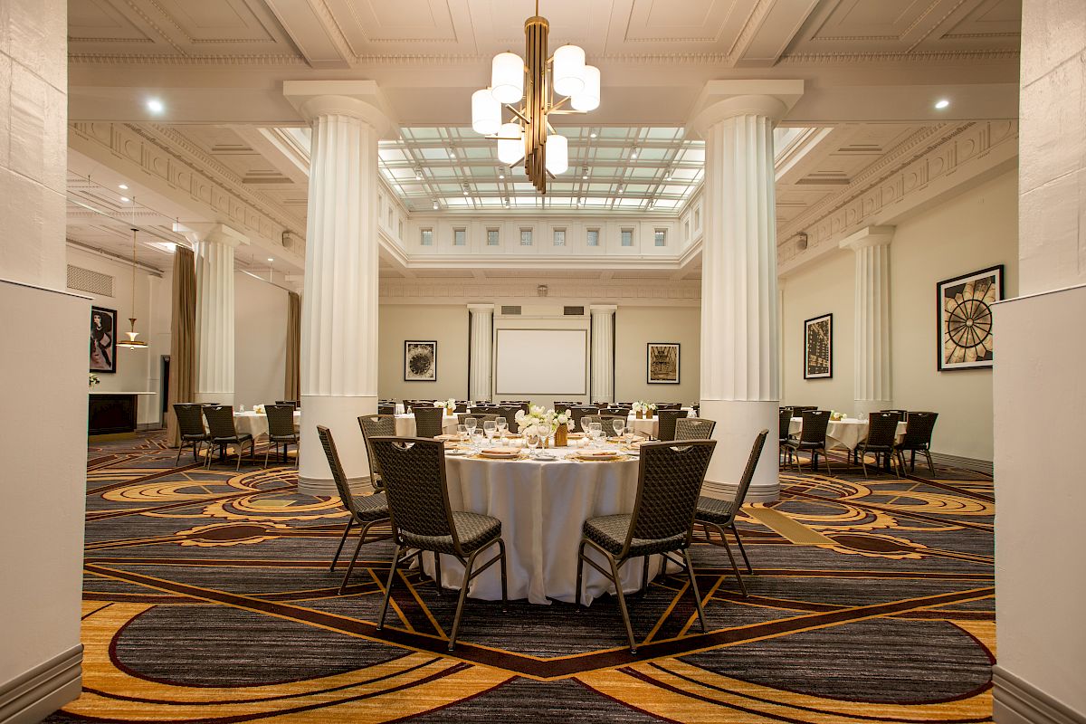 A spacious dining hall with round tables set for a formal event, featuring elegant lighting and decorative columns.