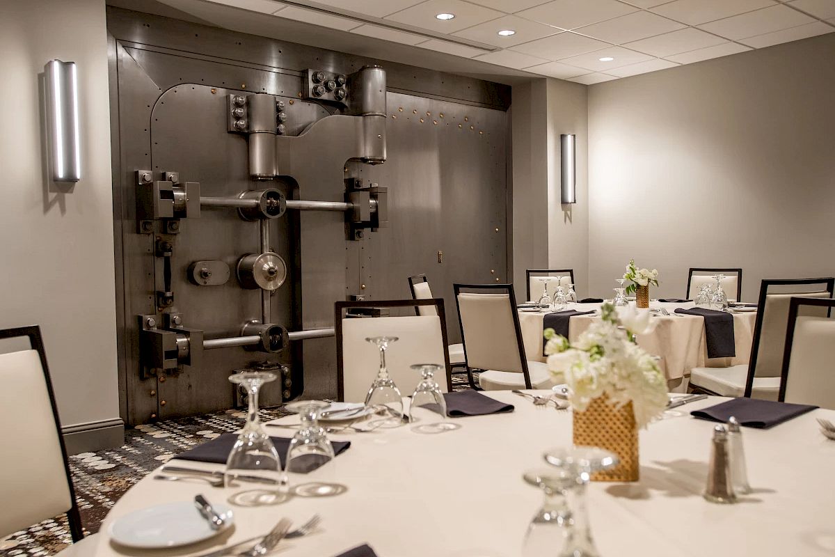 A dining area set against a large bank vault door, featuring elegantly arranged tables with glasses and napkins.