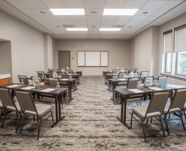 A modern conference room with arranged chairs and tables, notepads, and water glasses, facing a blank whiteboard, under ceiling lights.