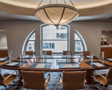 A modern conference room with a wooden table, leather chairs, notepads, and water bottles, set under a large chandelier.