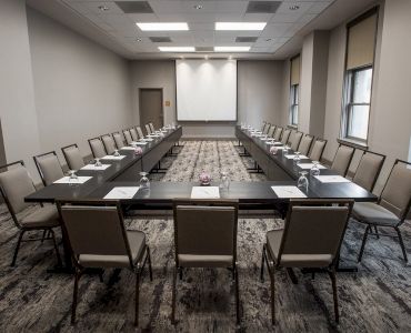A U-shaped conference room setup with chairs, tables, notepads, pens, and a projector screen, under fluorescent lighting, in a neutral-toned space.