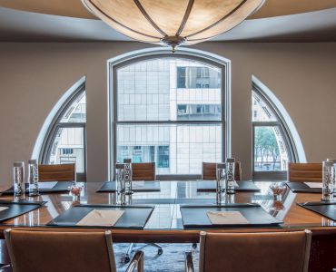 A modern conference room with a large table, chairs, notepads, water bottles, and an arched window view of a building outside.
