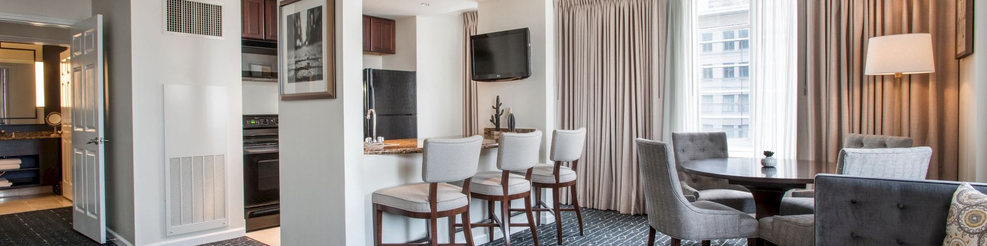 A modern hotel suite with a seating area, kitchenette, and bar stools, featuring a TV, large window, and contemporary decor.