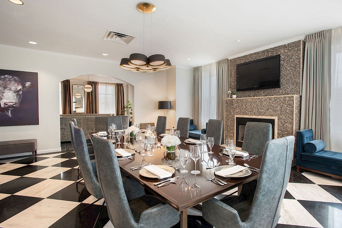 Elegant dining room with a set table, plush chairs, checkered floor, modern lighting, a painting, and a fireplace with a TV above it.