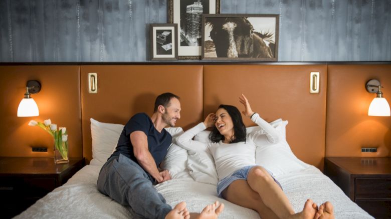 A couple is relaxing on a bed in a warmly lit room, with framed artwork above them and a vase of white flowers on the side table.