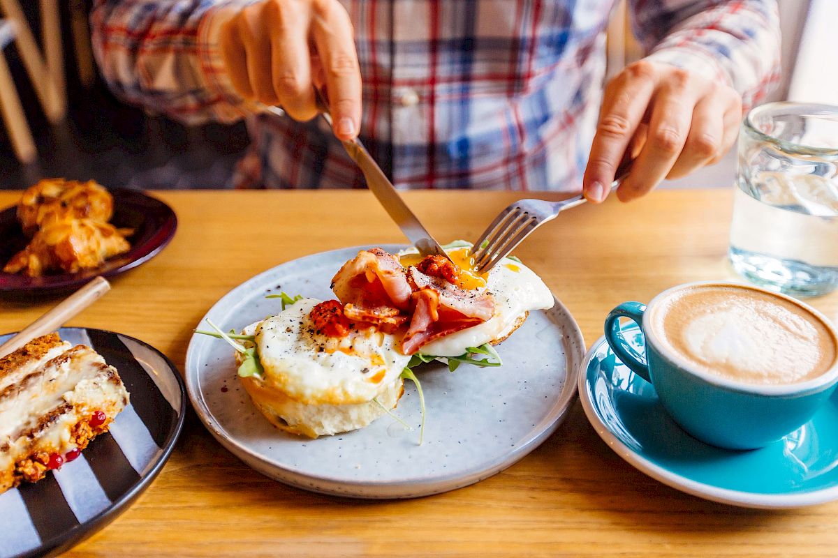 A person is eating a meal with eggs and bacon, accompanied by a cappuccino and dessert, seated at a wooden table with a glass of water.