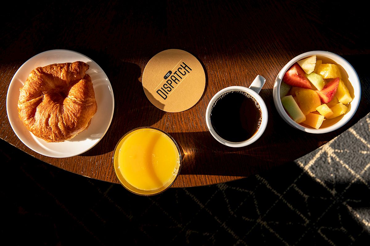 A breakfast setup with a croissant, orange juice, coffee, and a bowl of fruit on a table. A coaster with 
