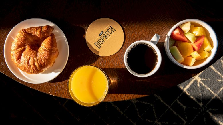 A breakfast setup with a croissant, orange juice, coffee, and a bowl of fruit on a table. A coaster with 