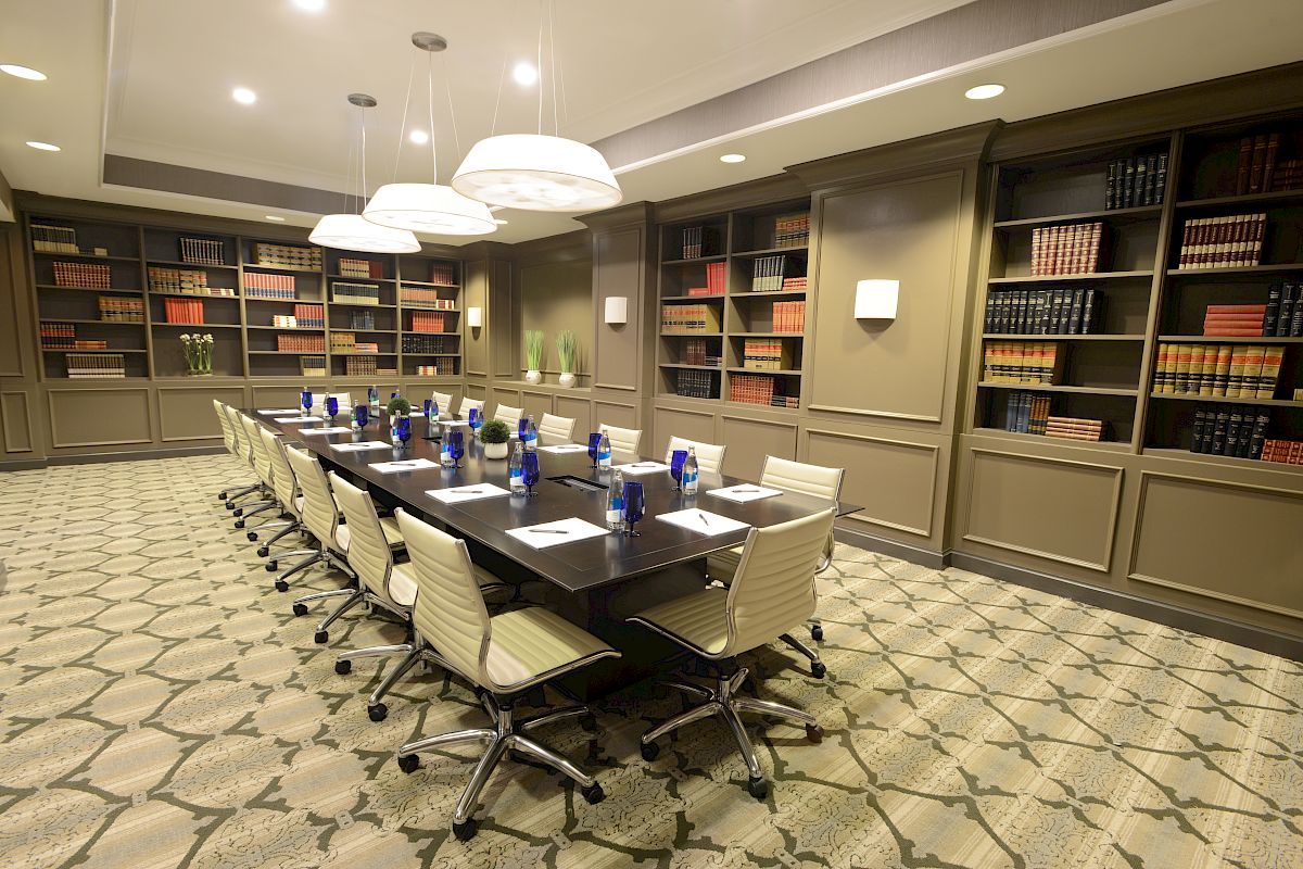 A conference room with a long table, surrounded by chairs, bookshelves, and notepads with water bottles on the table enhances the setting.
