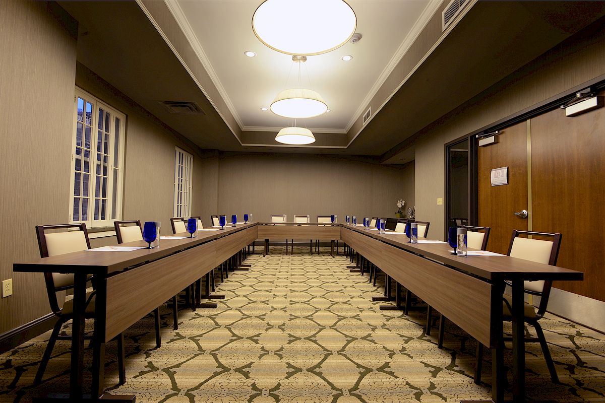 The image shows a conference room with U-shaped tables, chairs, and blue glasses, featuring overhead lighting and patterned carpet.