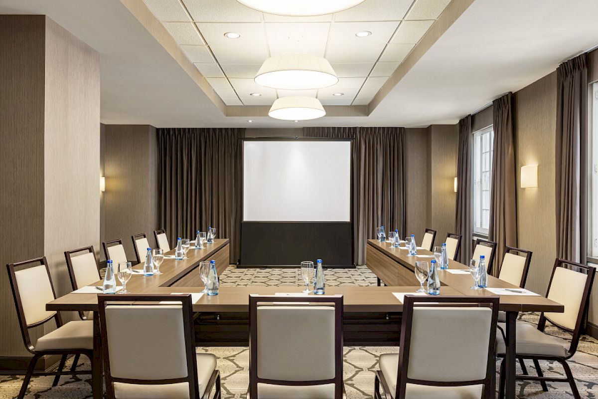 A conference room with a U-shaped table setup, chairs, a projector screen, water bottles, and note pads in a well-lit space.