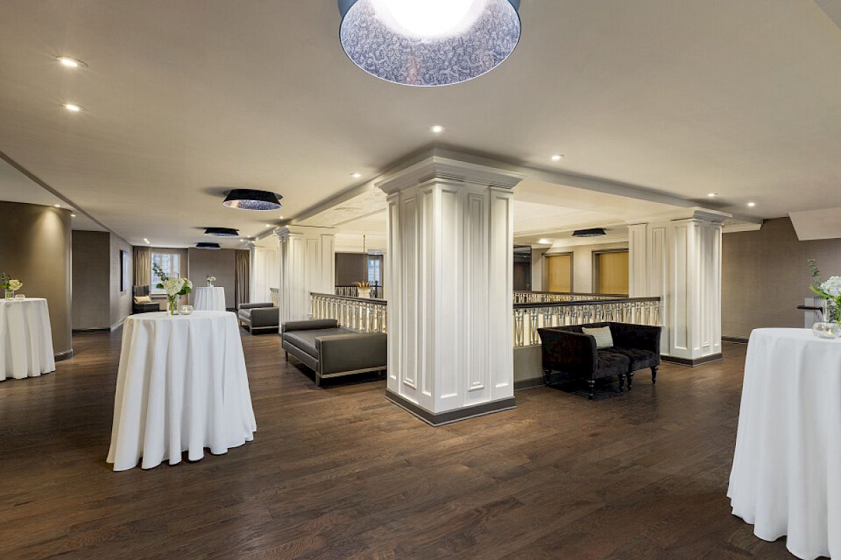 The image shows an elegant indoor space with white columns, round tables covered with cloths, and dark seating areas on wooden flooring.