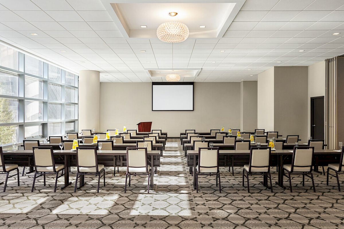 A conference room with rows of chairs, tables, a projector screen, and natural light from large windows, ready for a presentation.