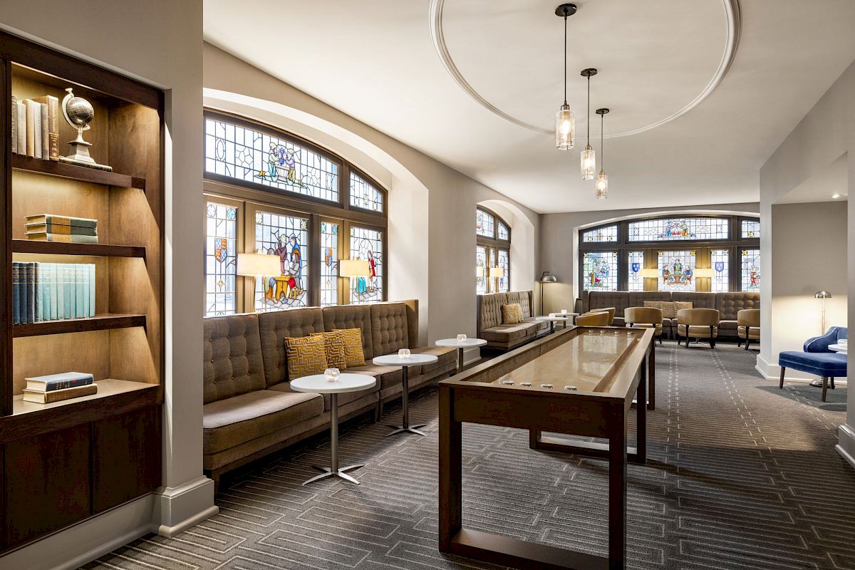 A cozy lounge with stained glass windows, sofa seating by the walls, a shuffleboard table, and bookshelves, creating a warm ambiance.