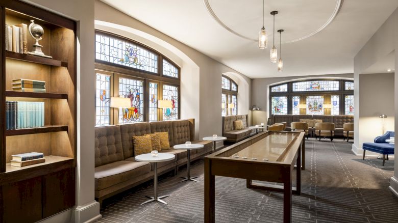 A cozy lounge with stained glass windows, sofa seating by the walls, a shuffleboard table, and bookshelves, creating a warm ambiance.