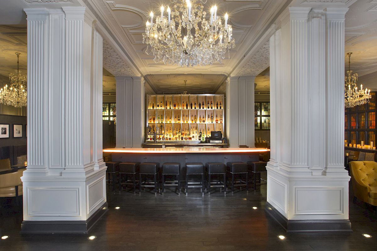 A modern bar with elegant chandeliers, tall white columns, and a dimly lit atmosphere, featuring a well-stocked backlit bar.
