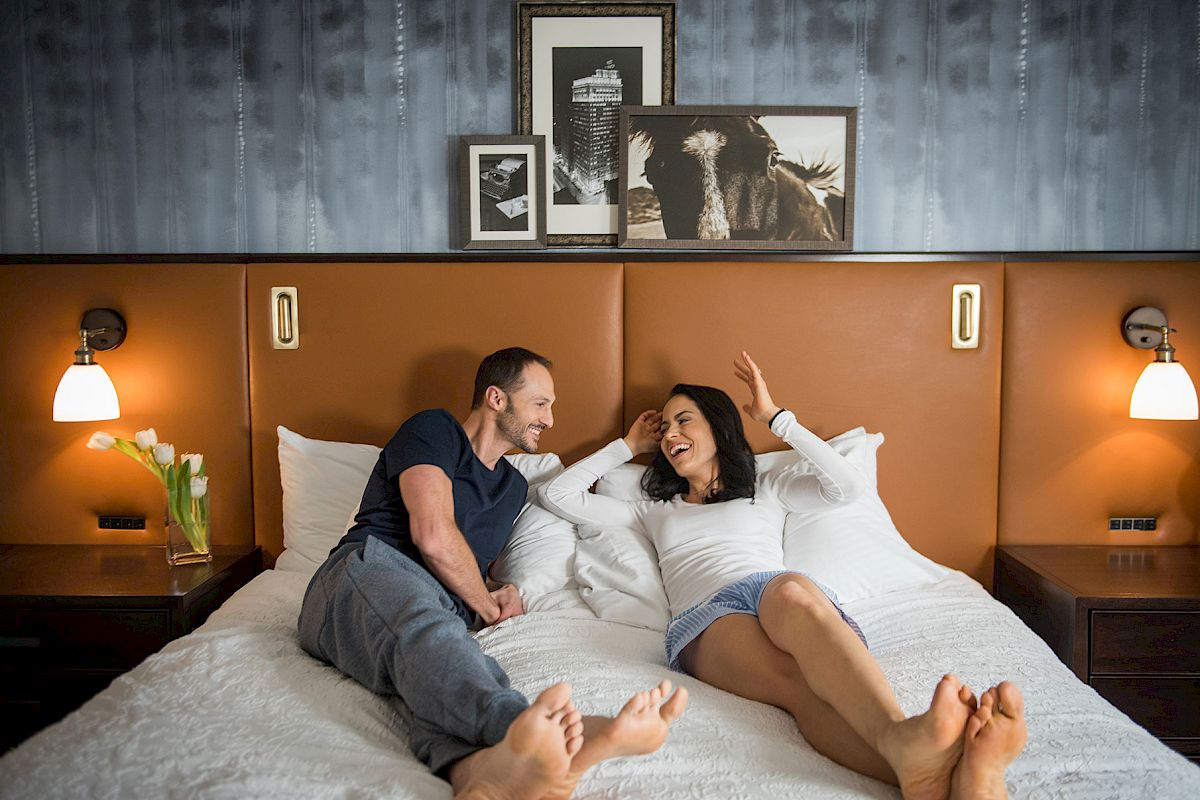 A couple is relaxing on a bed, smiling at each other in a warmly lit room with artwork and lamps on the wall.