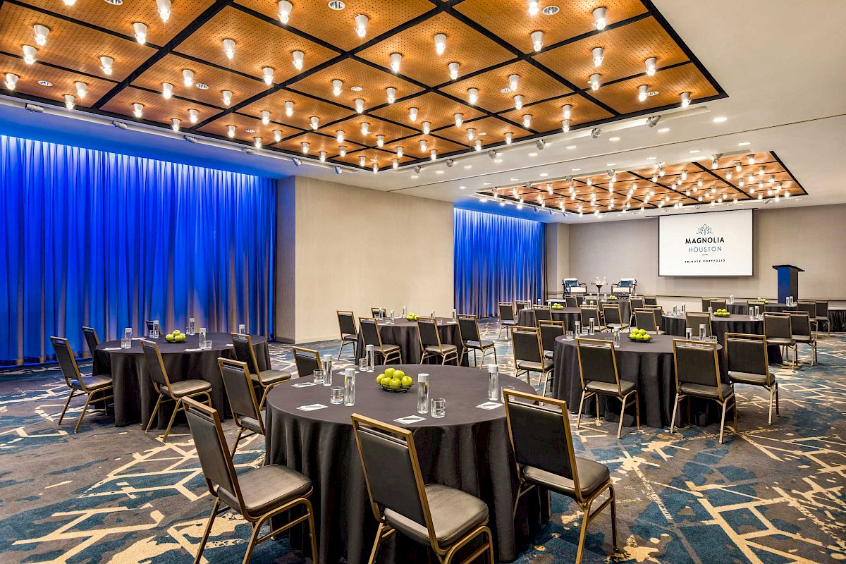 The image shows a modern conference room with round tables, chairs, a podium, overhead lights, and a screen displaying