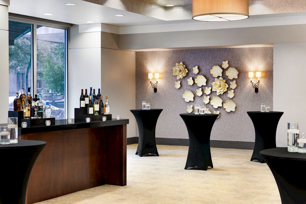 This image shows an elegant room setup for a reception with high-top tables, a bar with wine bottles, and floral wall decorations.