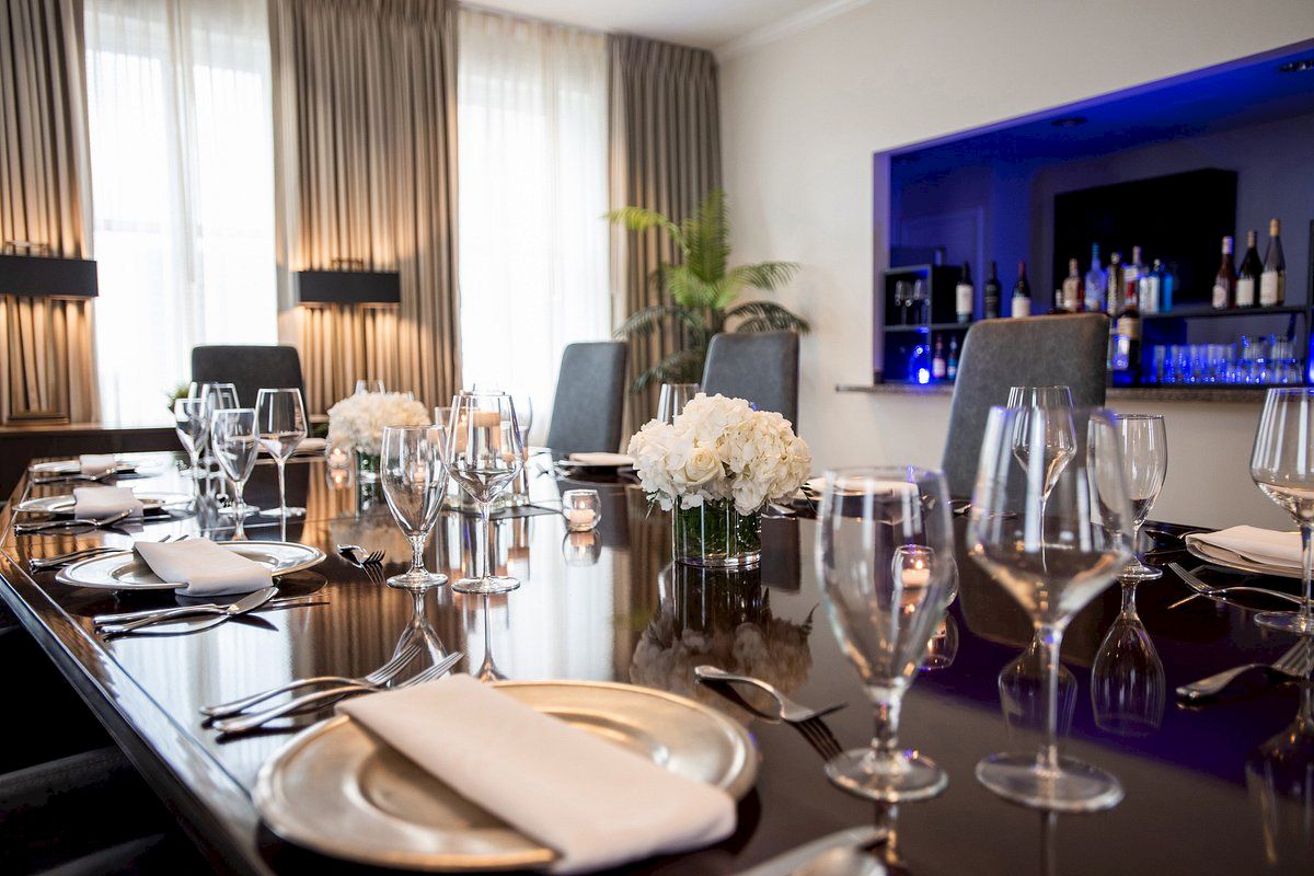 A dining table set elegantly for a formal gathering, with flowers, glassware, and napkins; a blue-lit bar is visible in the background.