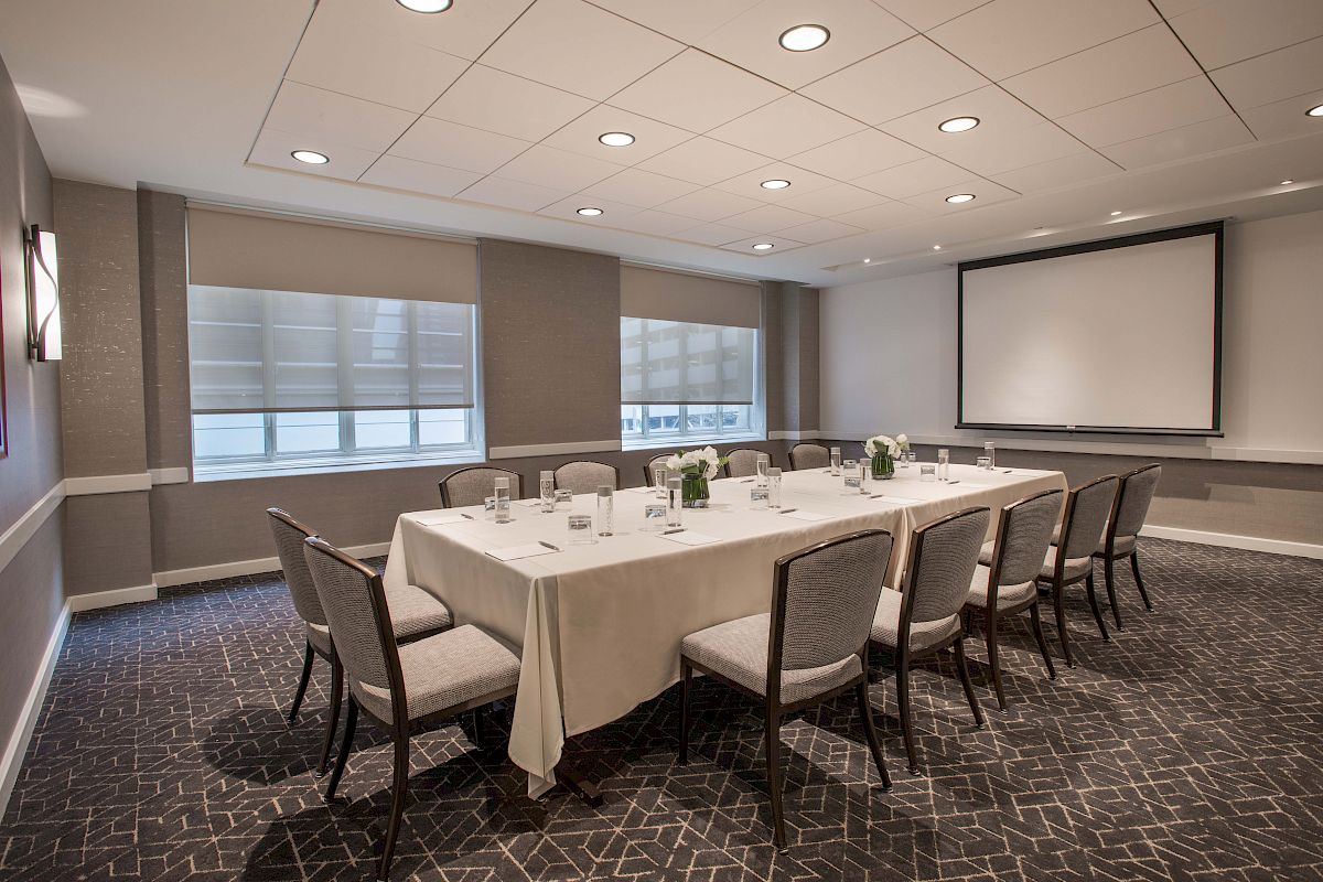 A conference room with a rectangular table, chairs, a projector screen, and a lit ceiling. The table is set with glasses and decorative items.