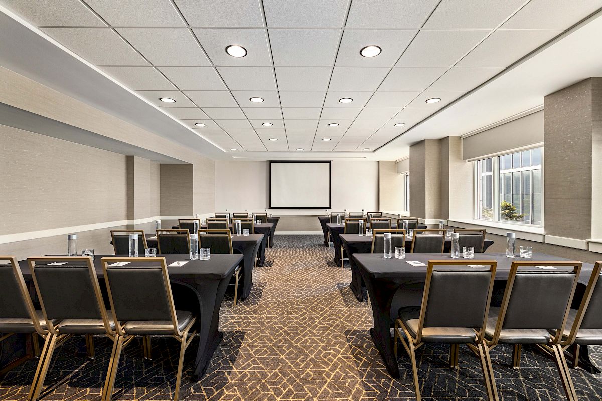A well-lit conference room with rows of tables and chairs facing a screen, ready for a meeting or presentation.