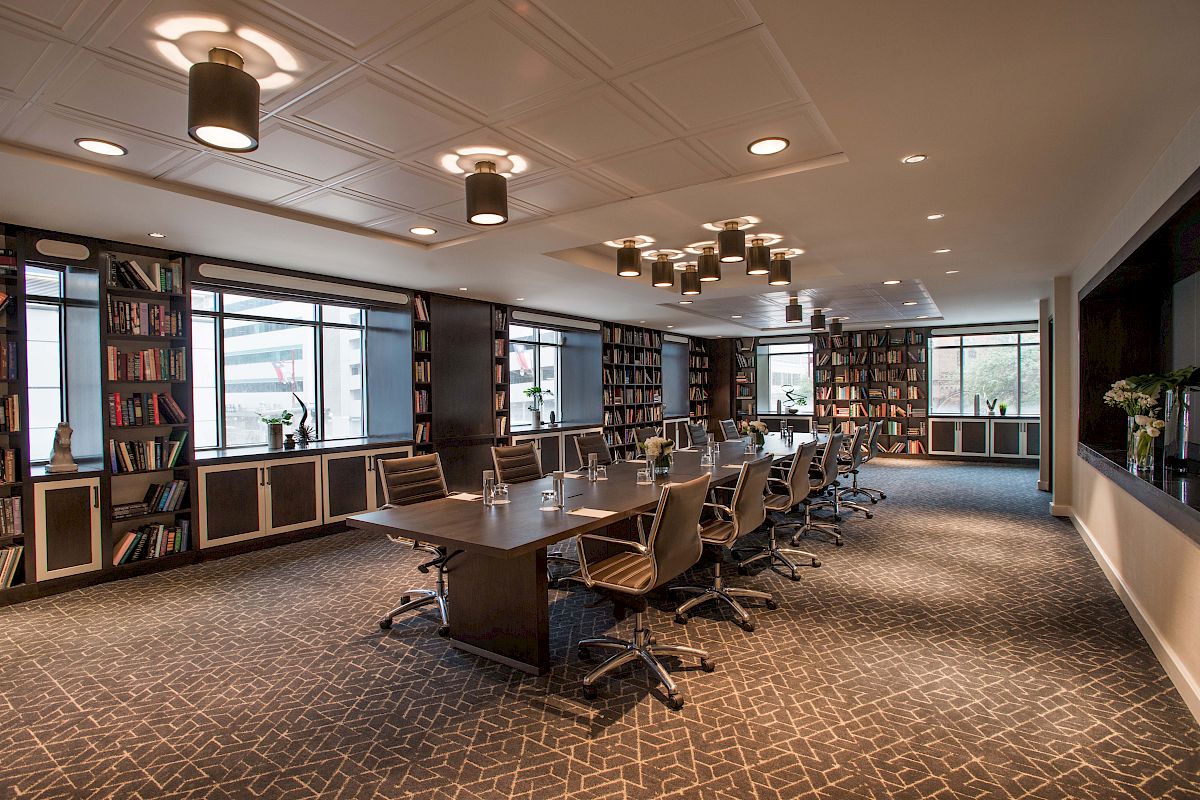 A modern conference room with a long table, chairs, bookshelves, and large windows, featuring overhead lighting for a professional setting.