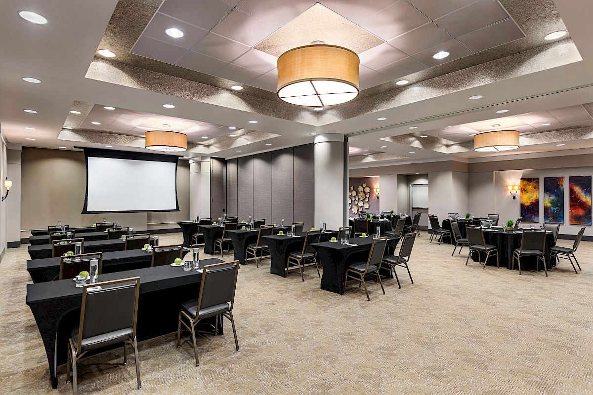 A conference room set up with tables and chairs facing a large screen, featuring modern lighting and abstract wall art on the sides.