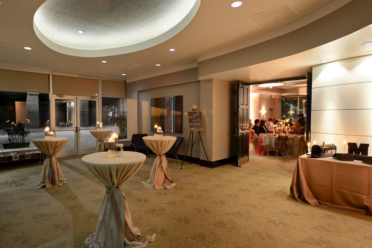 The image shows an elegantly decorated event space with round tables, draped in cloth, and a table in the back with a letter 