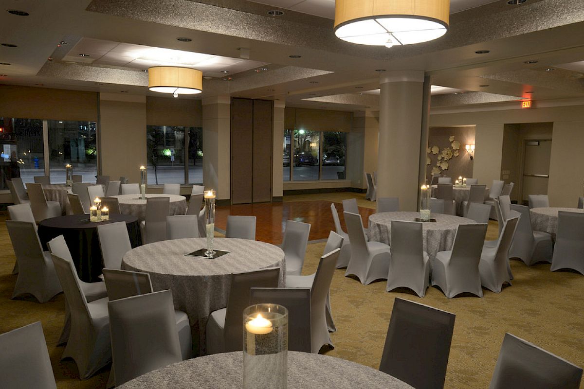 The image shows a well-lit, elegantly decorated banquet hall with round tables, grey covers, and candles on top.