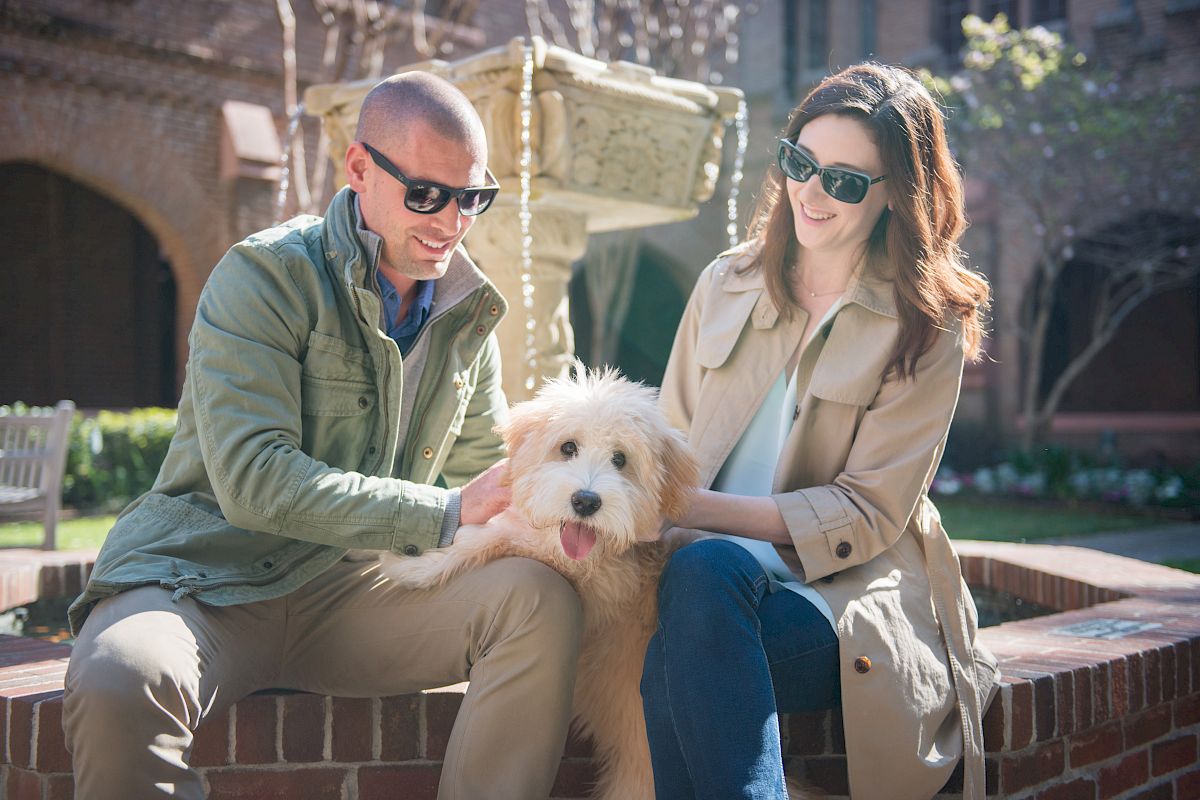 Two people wearing sunglasses sit by a fountain, smiling and petting a fluffy dog.