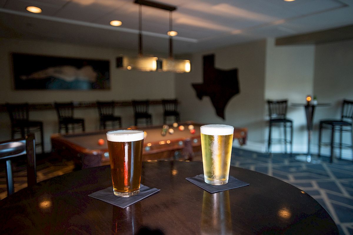 Two glasses of beer on a dark table in a dimly lit bar with Texas-shaped wall decor and seating in the background.