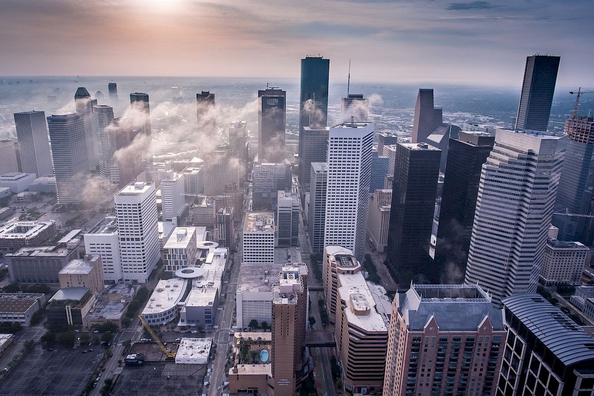 Aerial view of a cityscape with tall skyscrapers, clouds drifting between buildings, and a hazy sunlit sky.