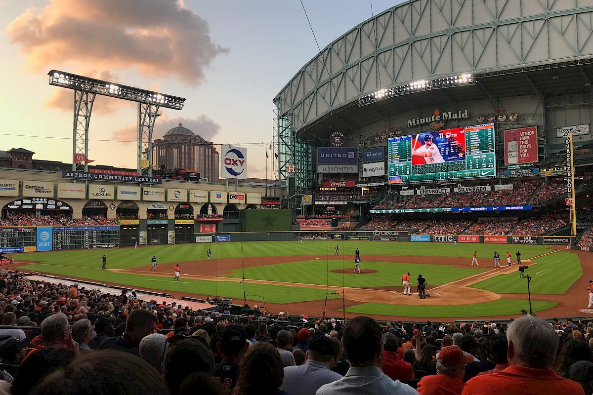 A baseball game is in progress at a large stadium, filled with spectators under a clouded sky as evening approaches.