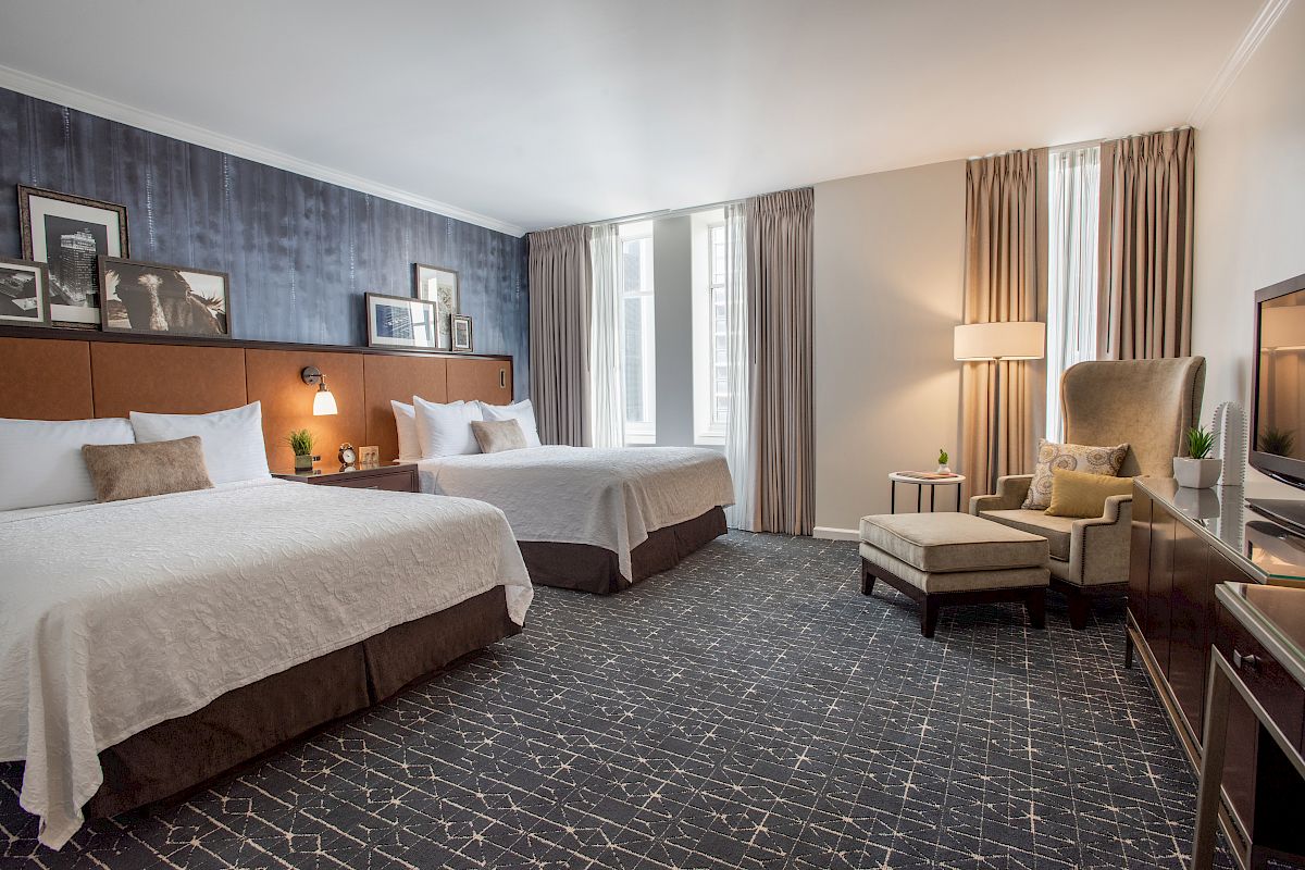 The image shows a hotel room with two beds, framed pictures, an armchair with a footrest, a floor lamp, and a TV, featuring large windows.