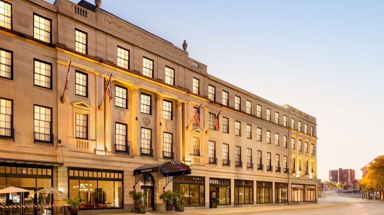 The image shows a grand, multi-story building with flags, large windows, and a welcoming entrance, likely a historic hotel or venue.