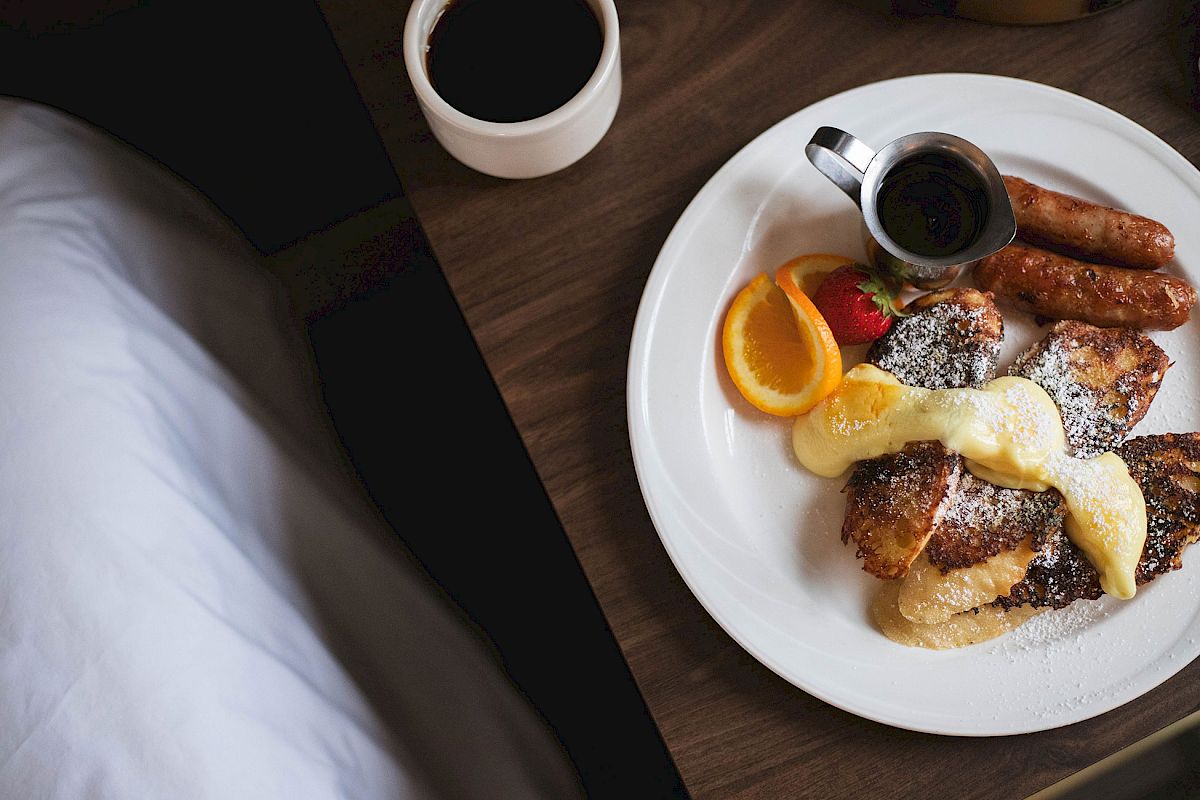 A breakfast plate with French toast, sausages, orange slice, strawberry, powdered sugar, and syrup; accompanied by a cup of coffee.