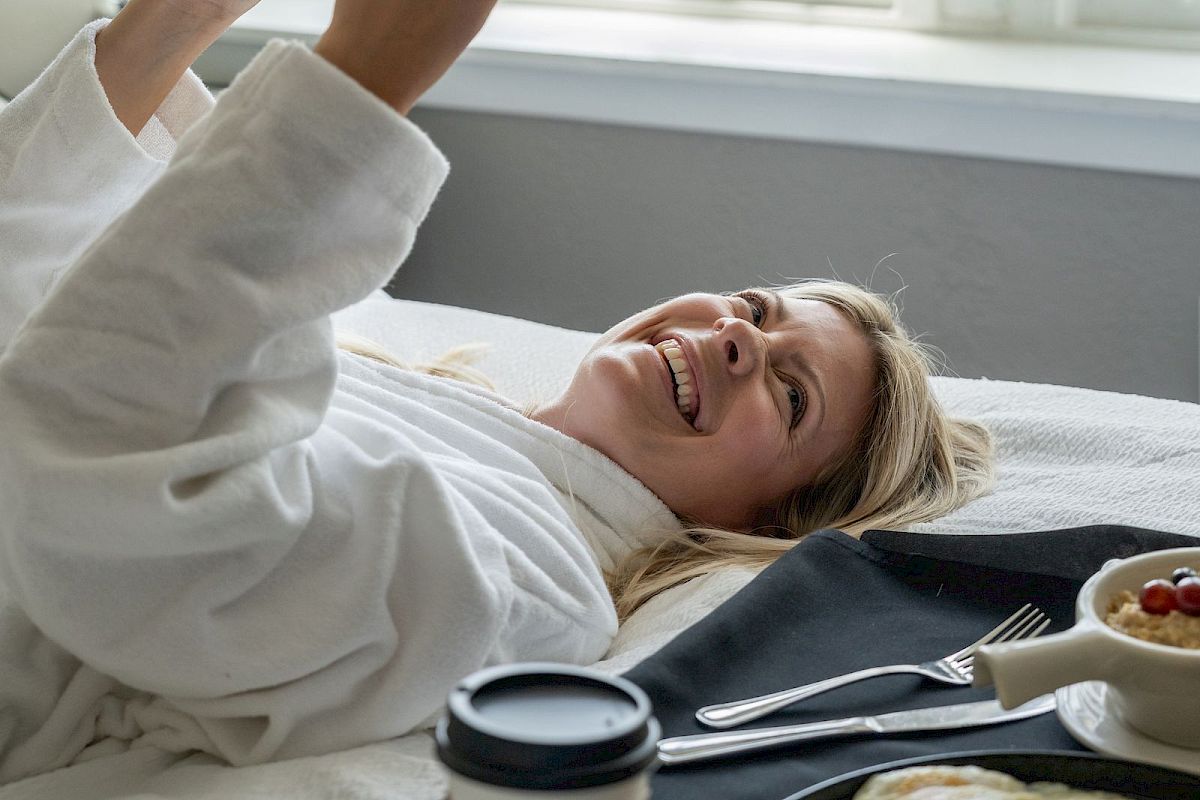 A person is lying on a bed reading a book, with breakfast and a coffee on a tray beside them.